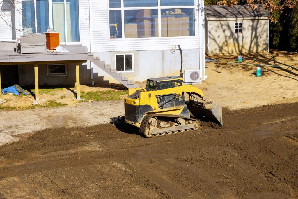 Skid steer grading soil near the foundation of a house to create proper slope and drainage.
